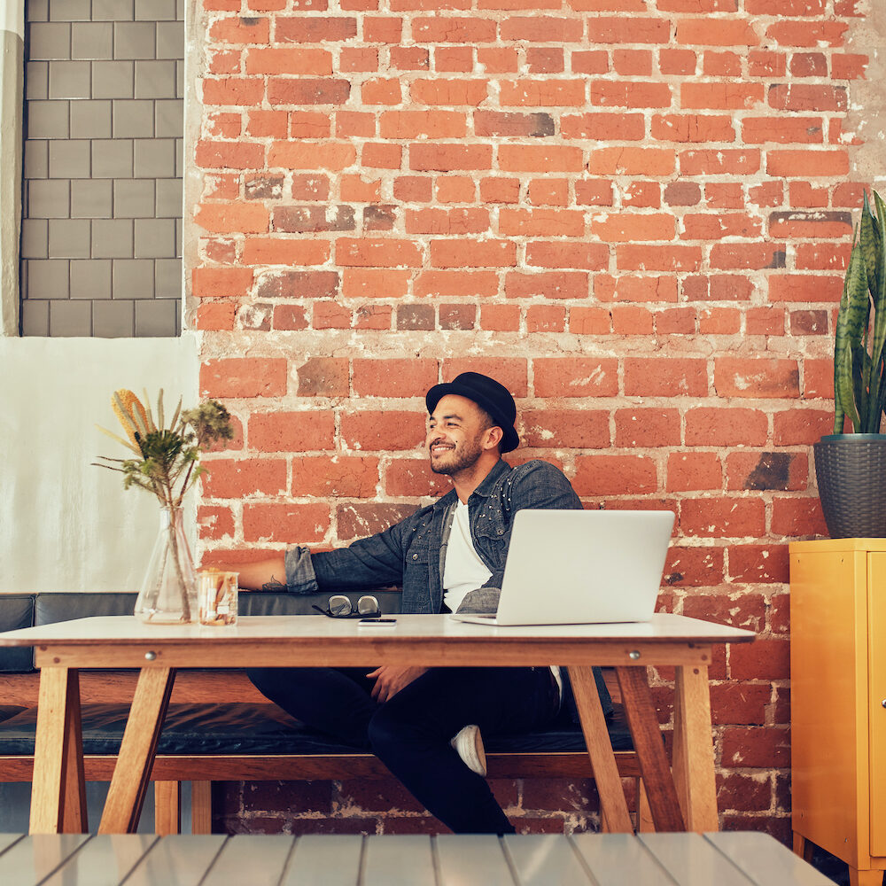 Portrait of happy young man sitting at a cafe with laptop on table. Caucasian man at coffee shop looking away and smiling.