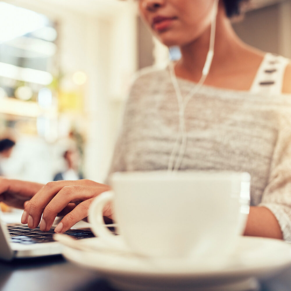 Woman sitting at a coffee shop working on laptop, with focus on hands typing on keyboard.